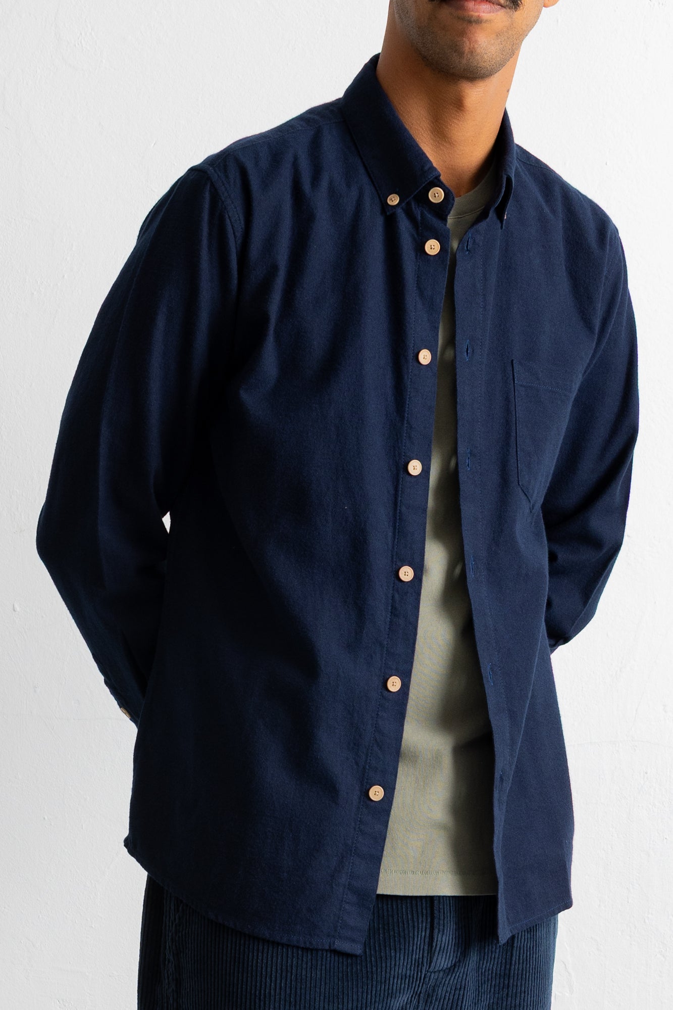 A man wearing a navy blue long-sleeve button-down shirt with a chest pocket, layered over a light olive green t-shirt, against a plain white background.