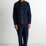 A man wearing a dark navy blue long-sleeve button-up shirt, matching navy pants, and black canvas sneakers, standing against a white studio wall.