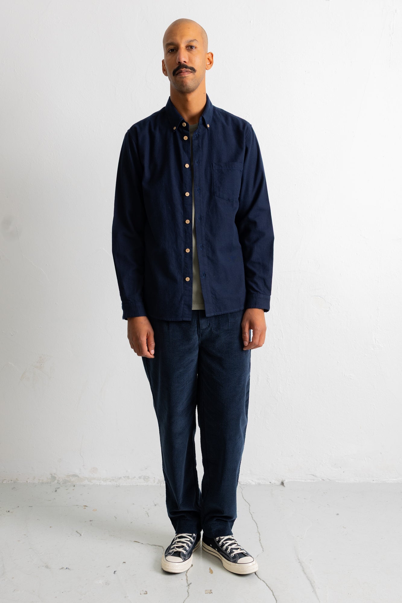 A man wearing a dark navy blue long-sleeve button-up shirt, matching navy pants, and black canvas sneakers, standing against a white studio wall.