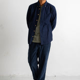 A man wearing a navy blue button-up overshirt with gold-tone buttons, dark navy pants, a muted green t-shirt, and black canvas sneakers, standing against a white studio wall.
