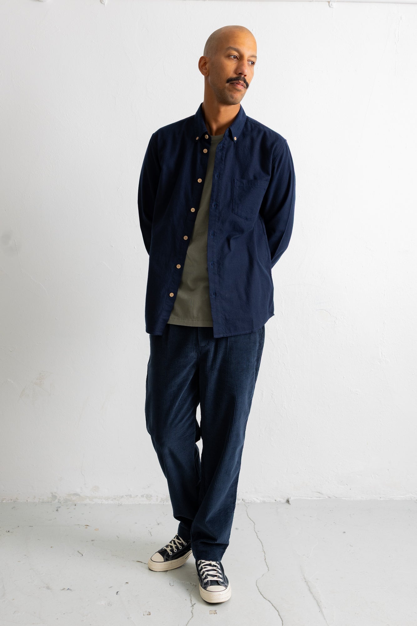 A man wearing a navy blue button-up overshirt with gold-tone buttons, dark navy pants, a muted green t-shirt, and black canvas sneakers, standing against a white studio wall.