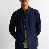 A man wearing a dark navy blue corduroy button-down shirt over a light olive green t-shirt and dark corduroy trousers, standing against a plain white wall.