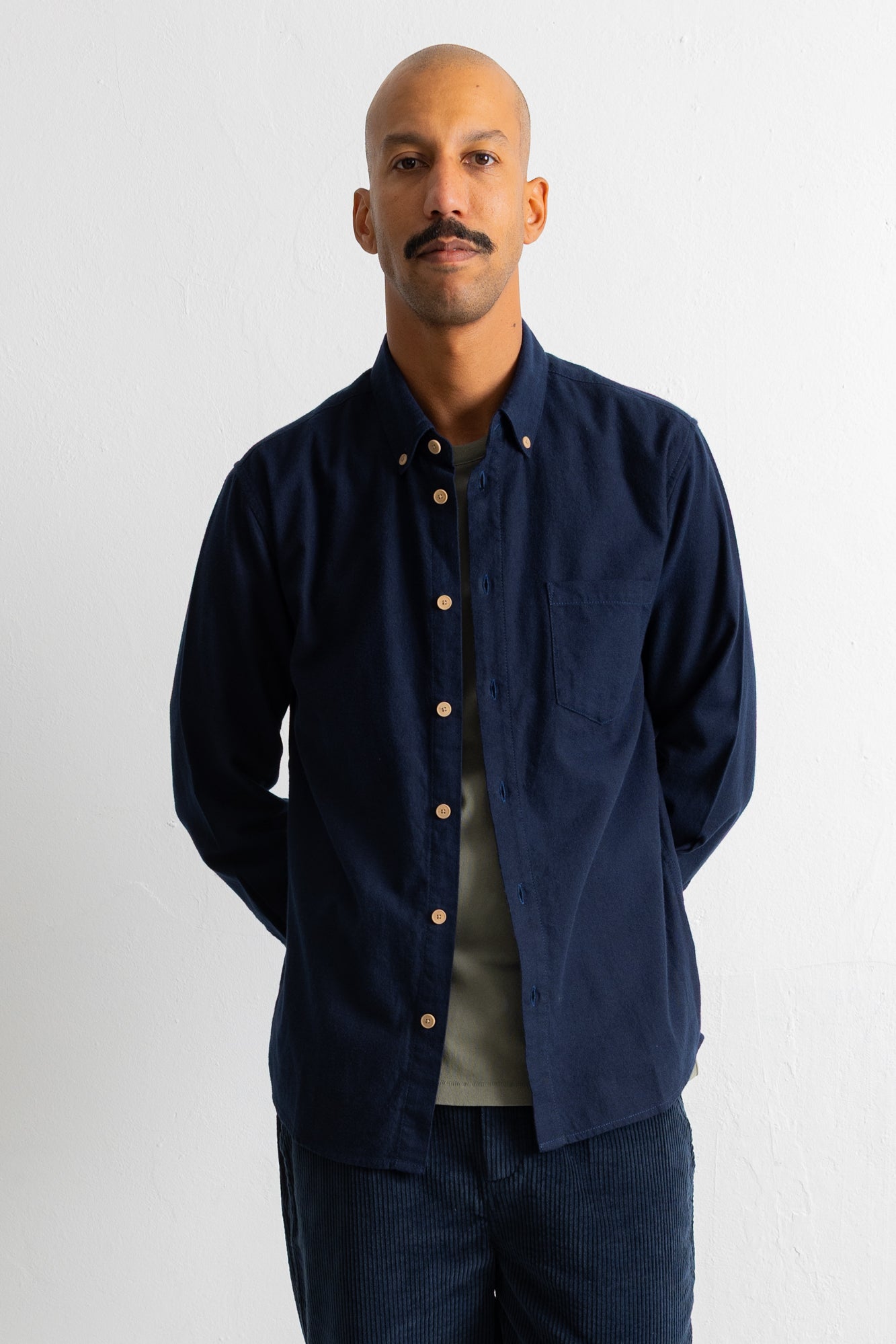 A man wearing a dark navy blue corduroy button-down shirt over a light olive green t-shirt and dark corduroy trousers, standing against a plain white wall.