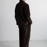 A person standing in profile wearing a dark brown corduroy jacket and matching wide-leg trousers against a plain white studio wall.