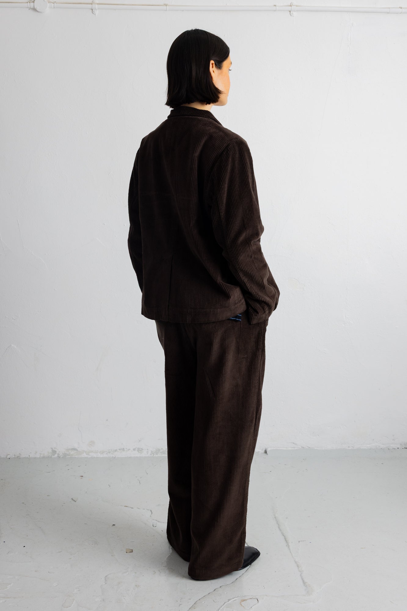 A person standing in profile wearing a dark brown corduroy jacket and matching wide-leg trousers against a plain white studio wall.