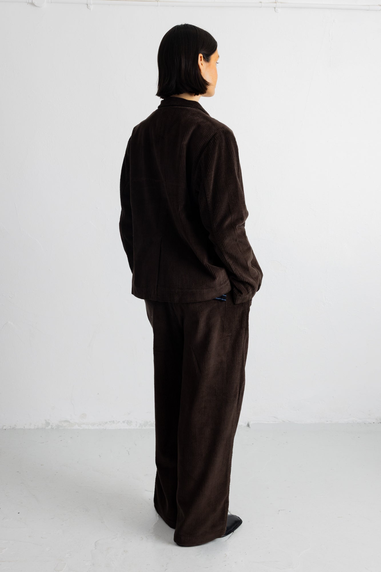 A model shown from the back wearing a dark brown corduroy jacket and matching wide-leg trousers, standing against a plain white studio background.