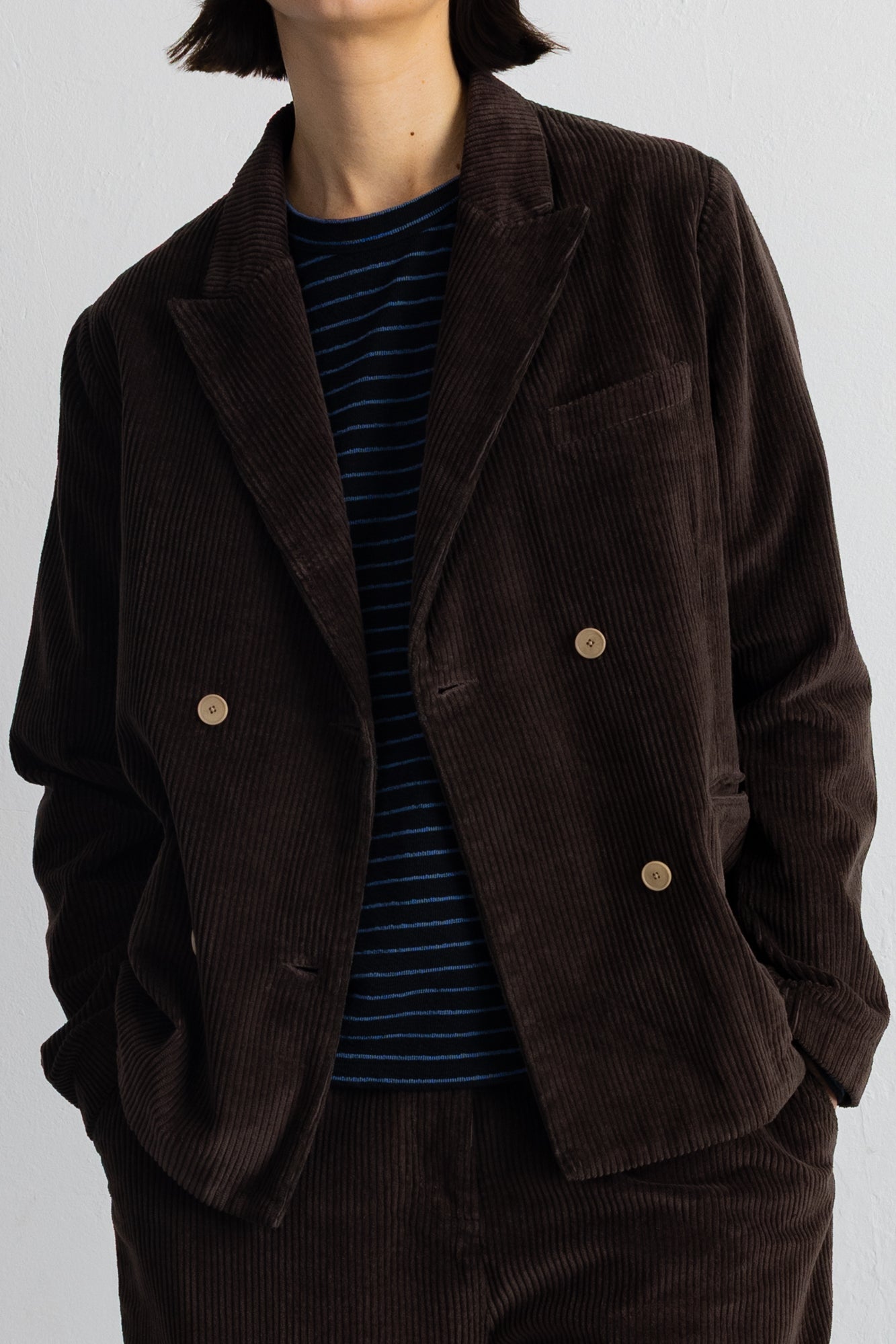 A person wearing a dark brown corduroy blazer over a navy and blue striped shirt, paired with matching corduroy trousers, against a plain light gray background.