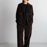 A model stands against a light gray wall wearing a dark brown corduroy double-breasted blazer and matching wide-leg trousers over a navy and white striped shirt, paired with black shoes.