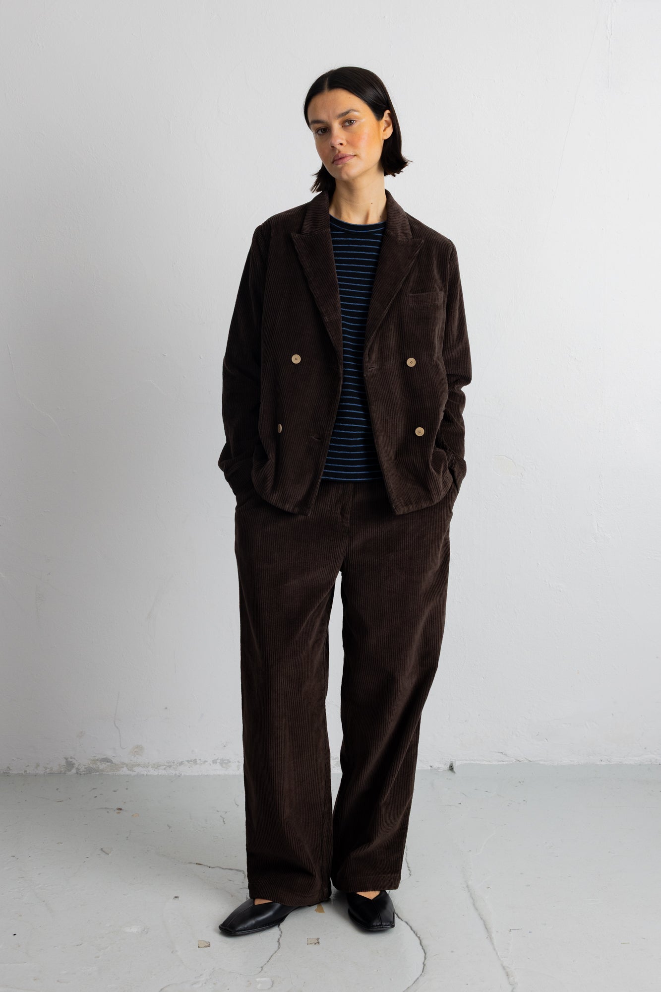 A model stands against a light gray wall wearing a dark brown corduroy double-breasted blazer and matching wide-leg trousers over a navy and white striped shirt, paired with black shoes.
