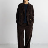 A model stands against a plain white studio wall wearing a dark brown corduroy double-breasted jacket with gold buttons, matching wide-leg trousers, a navy and black striped shirt, and black shoes.