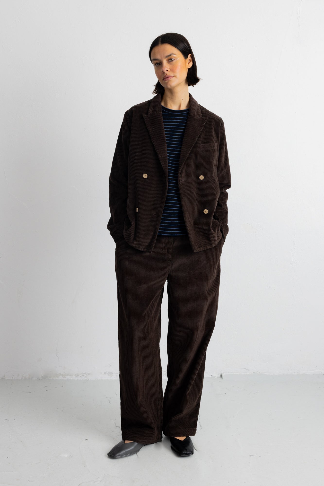 A model stands against a plain white studio wall wearing a dark brown corduroy double-breasted jacket with gold buttons, matching wide-leg trousers, a navy and black striped shirt, and black shoes.