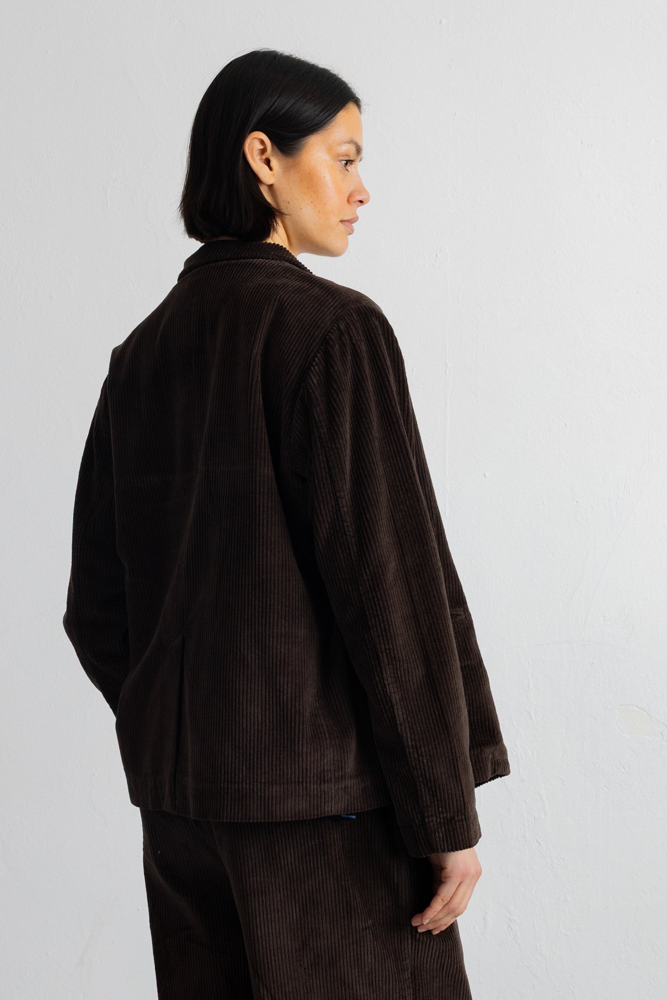 A model shown from the back wearing a dark brown corduroy jacket and matching corduroy trousers against a plain light gray background.