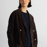 A model wearing a dark brown corduroy double-breasted blazer over a navy and blue striped t-shirt, paired with matching corduroy trousers, shot against a plain light gray background.