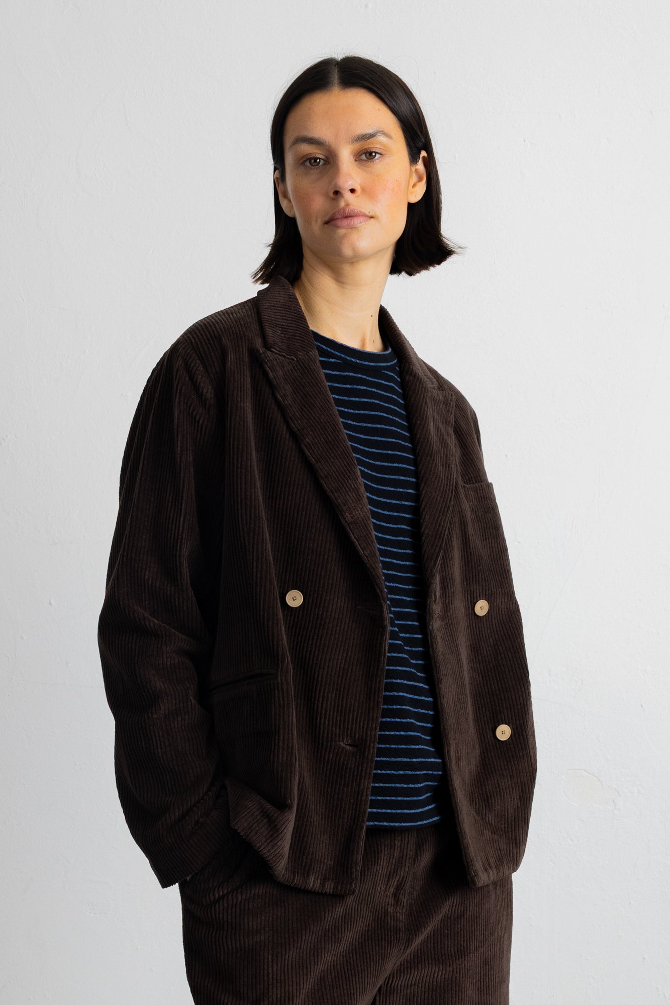 A model wearing a dark brown corduroy double-breasted blazer over a navy and blue striped t-shirt, paired with matching corduroy trousers, shot against a plain light gray background.