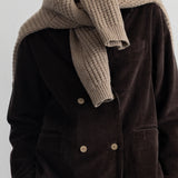 A close-up of a person wearing a thick, ribbed knit scarf in a light taupe-beige color, styled over a dark brown corduroy jacket.