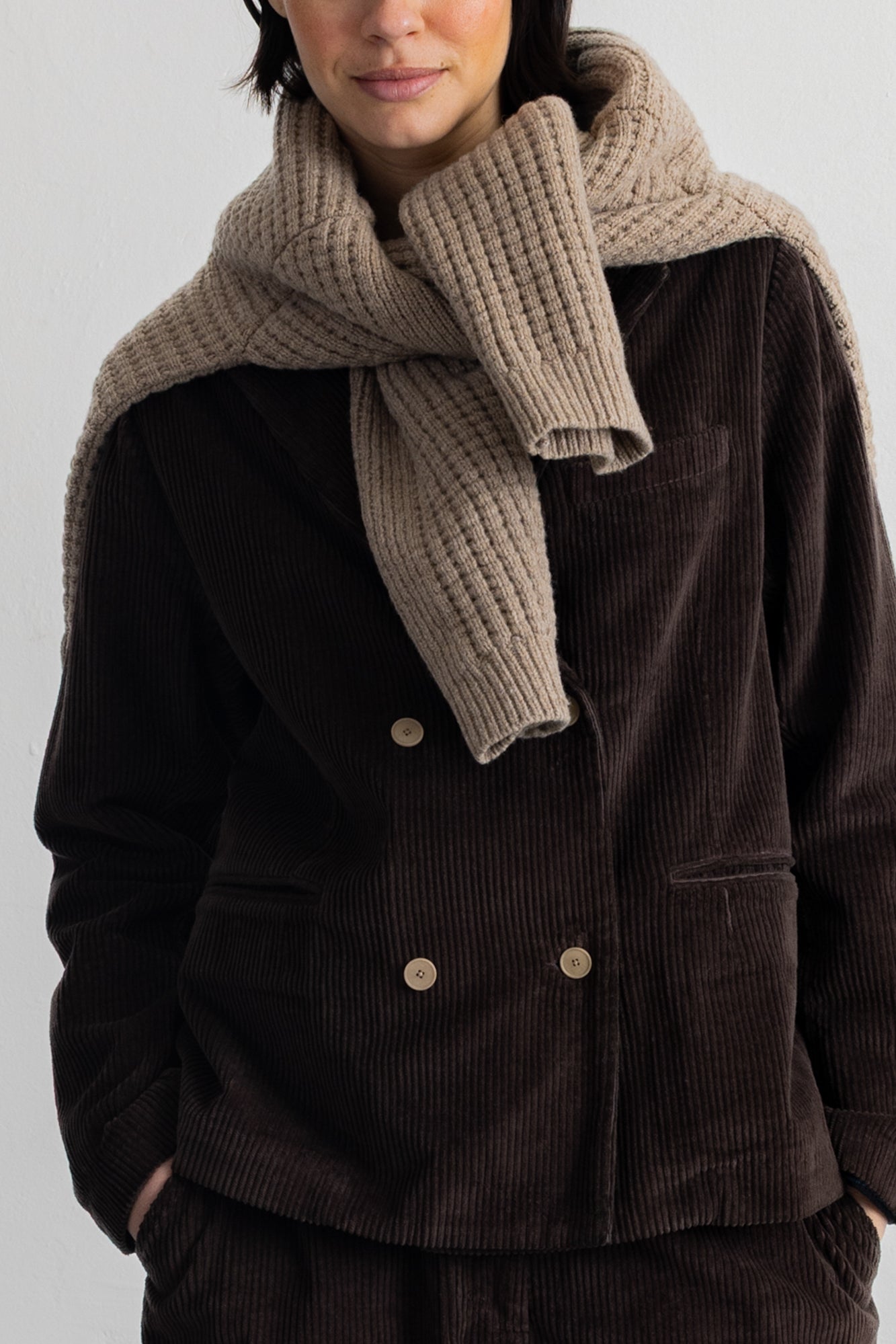 A close-up of a person wearing a thick, ribbed knit scarf in a light taupe-beige color, styled over a dark brown corduroy jacket.