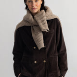A woman wearing a dark brown corduroy jacket and matching corduroy pants, styled with a chunky beige knit scarf, against a plain light gray background.