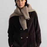 A model stands against a plain white studio wall wearing a dark brown corduroy double-breasted jacket with gold buttons, matching wide-leg trousers, a navy and black striped shirt, and black shoes.
