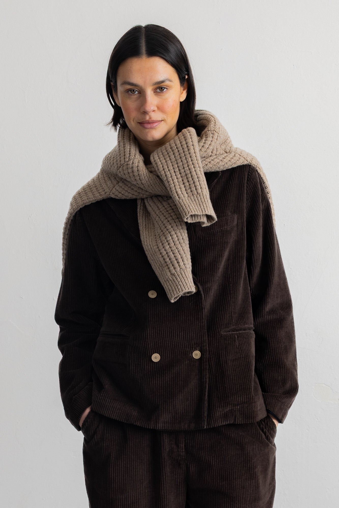 A model stands against a plain white studio wall wearing a dark brown corduroy double-breasted jacket with gold buttons, matching wide-leg trousers, a navy and black striped shirt, and black shoes.
