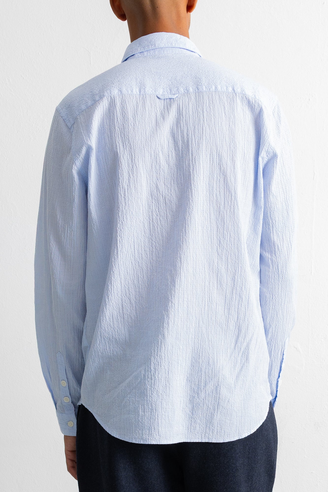 A person shown from the back wearing a light blue textured long-sleeve button-up shirt and dark trousers against a plain white background.
