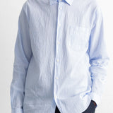 A man wearing a light blue long-sleeve button-down shirt with a textured crinkle fabric, paired with dark trousers, against a white wall.