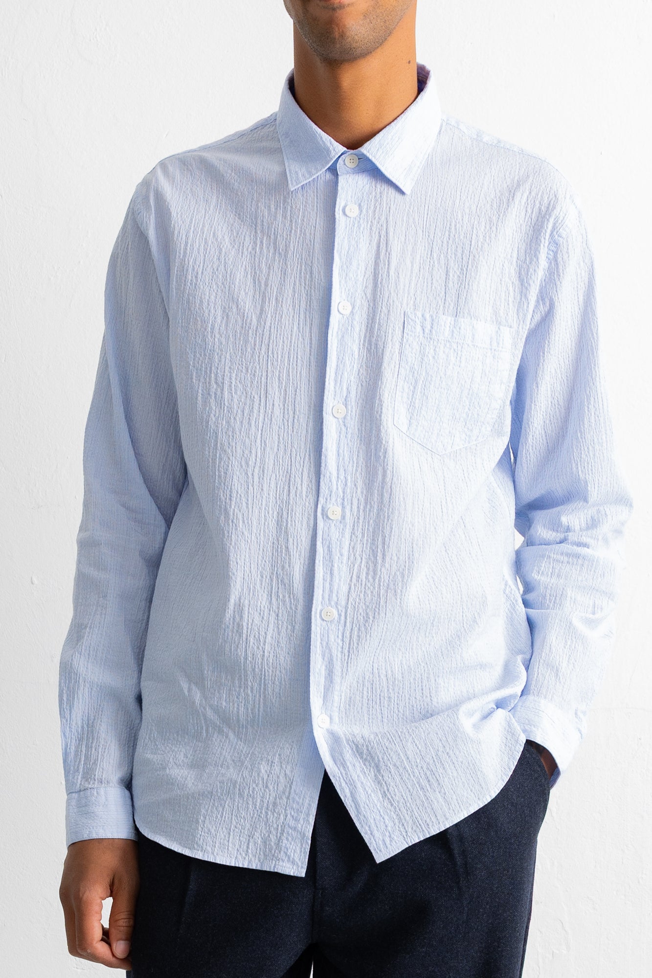 A man wearing a light blue long-sleeve button-down shirt with a textured crinkle fabric, paired with dark trousers, against a white wall.