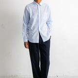A man wearing a light blue and white striped long-sleeve button-down shirt, dark navy trousers, and dark brown leather dress shoes, standing against a white wall.