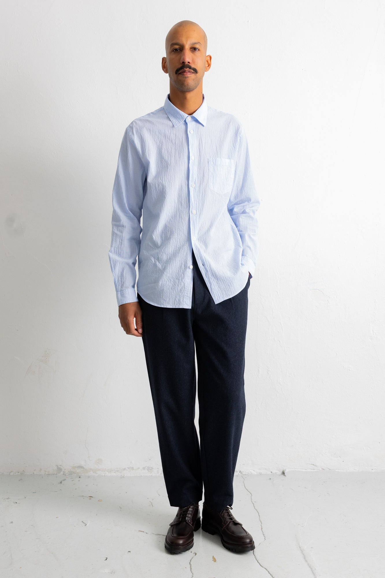 A man wearing a light blue and white striped long-sleeve button-down shirt, dark navy trousers, and dark brown leather dress shoes, standing against a white wall.