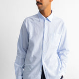A man wearing a light blue and white vertically striped long-sleeve button-down shirt, paired with dark trousers, standing against a plain white wall.