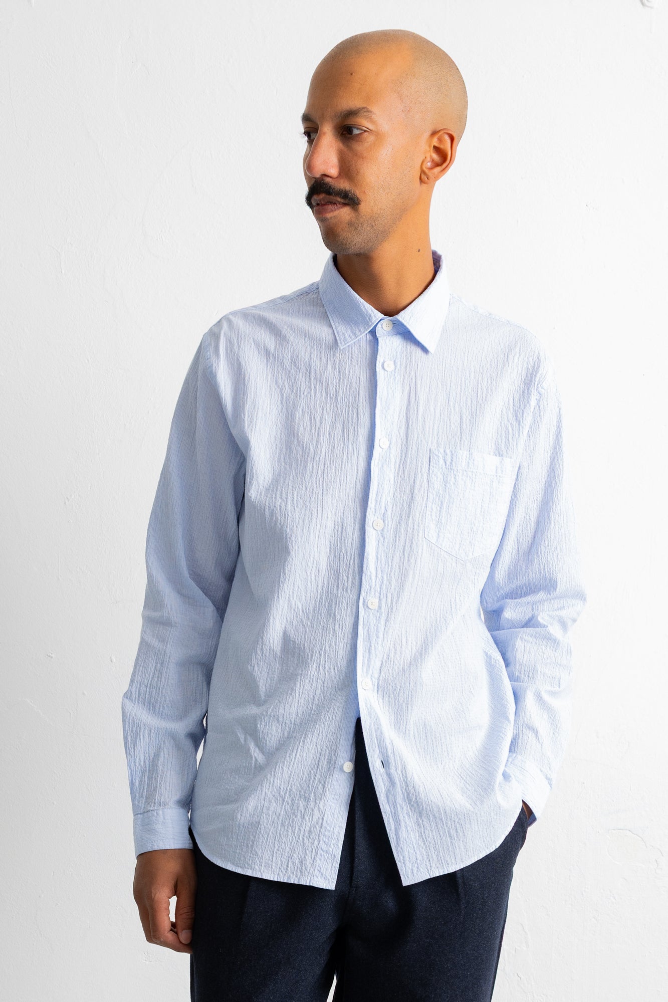 A man wearing a light blue and white vertically striped long-sleeve button-down shirt, paired with dark trousers, standing against a plain white wall.