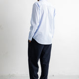 A man standing with his back to the camera, wearing a light blue and white striped long-sleeve button-down shirt, dark navy trousers, and dark brown leather shoes in a minimalist white room.