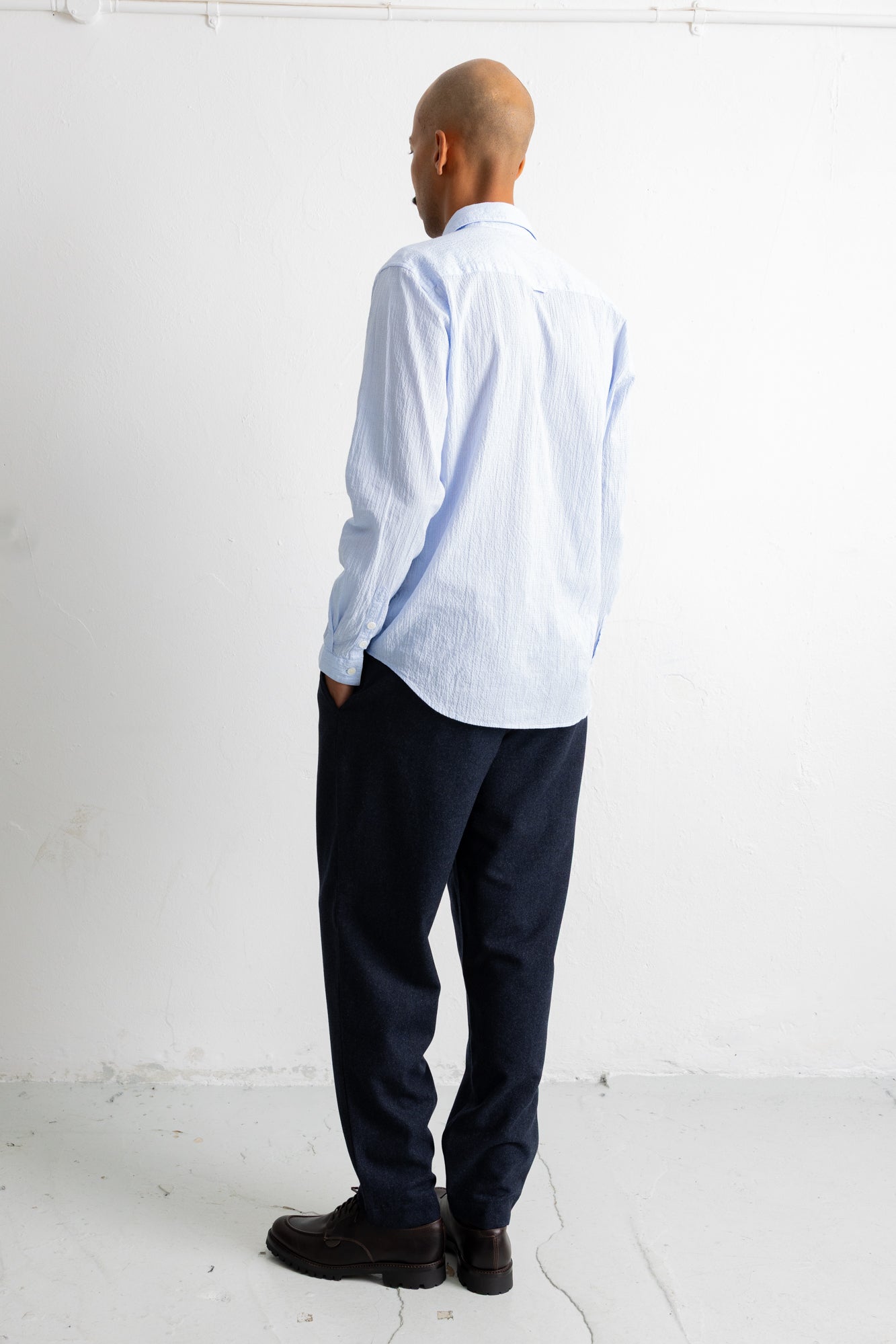 A man standing with his back to the camera, wearing a light blue and white striped long-sleeve button-down shirt, dark navy trousers, and dark brown leather shoes in a minimalist white room.