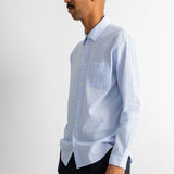 A man in profile wearing a light blue long-sleeve button-down shirt with a chest pocket and dark trousers, standing against a plain off-white wall.