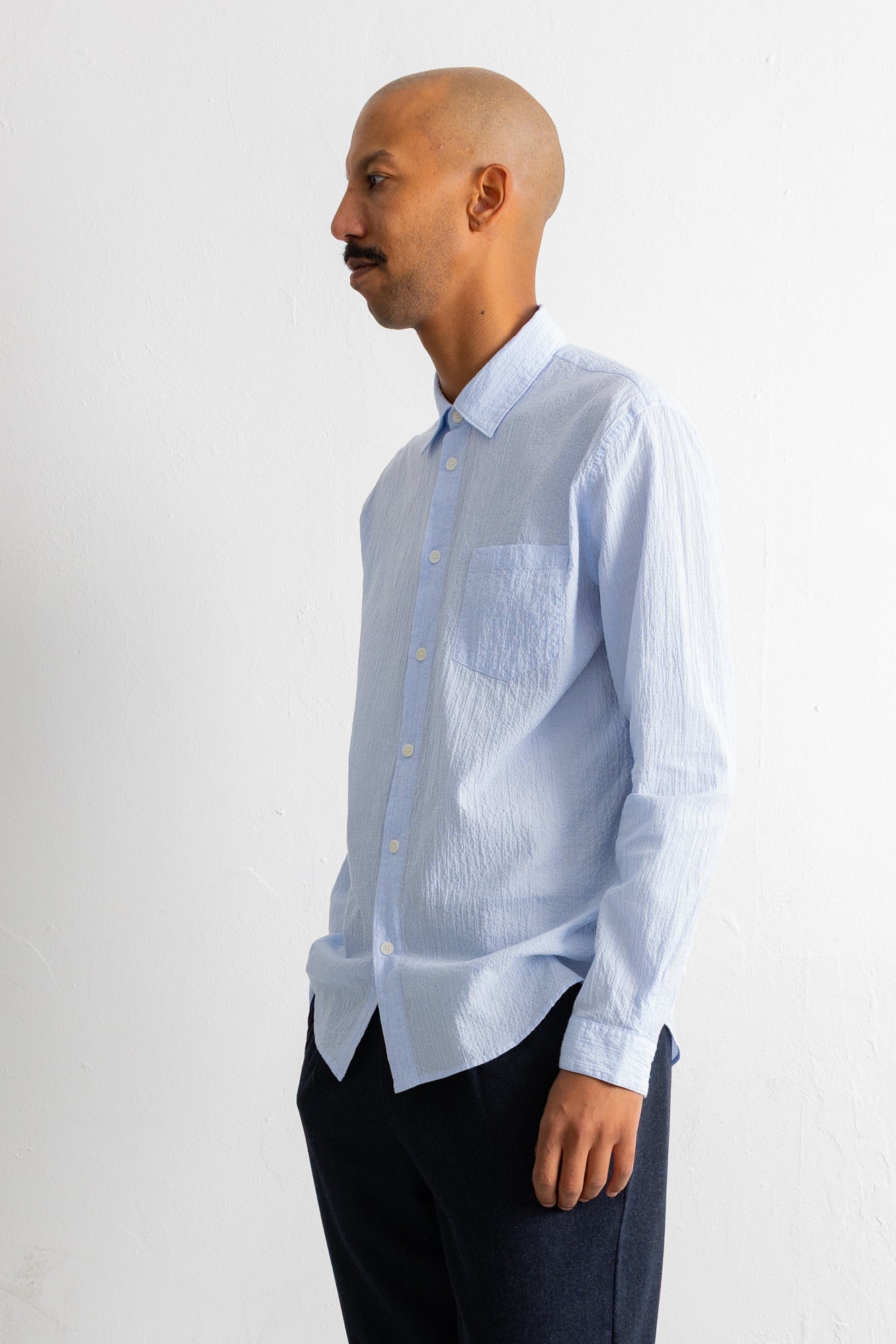 A man in profile wearing a light blue long-sleeve button-down shirt with a chest pocket and dark trousers, standing against a plain off-white wall.