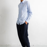 A man wearing a light blue long-sleeve button-down shirt, dark navy trousers, and dark brown leather dress shoes, standing in profile against a white wall.