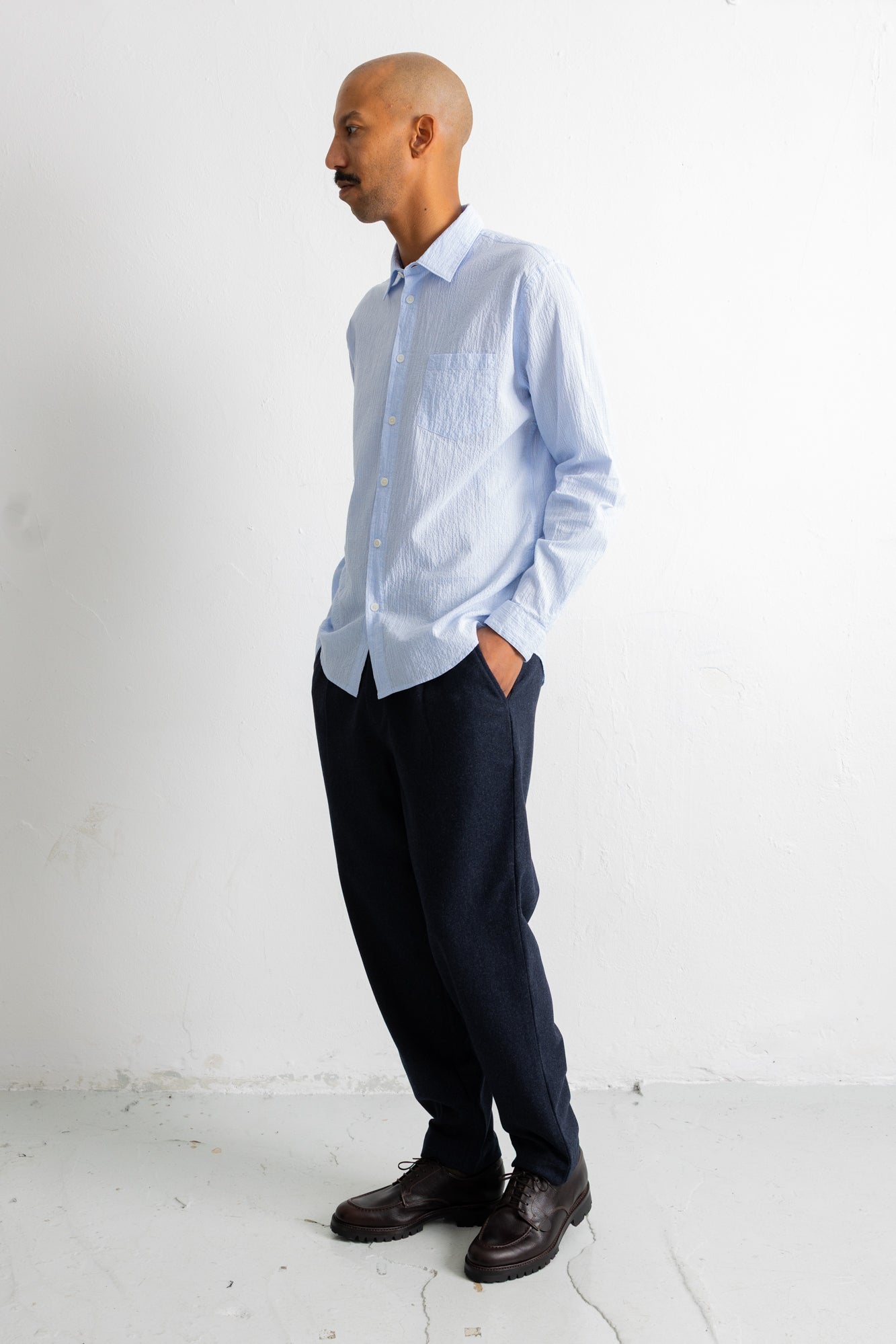 A man wearing a light blue long-sleeve button-down shirt, dark navy trousers, and dark brown leather dress shoes, standing in profile against a white wall.