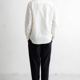 A man viewed from behind wearing a white textured long-sleeve button-up shirt, dark trousers, and black shoes, standing against a plain white wall.
