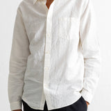 A man wearing a long-sleeve white textured linen button-down shirt with a chest pocket, paired with dark trousers, against a white wall.