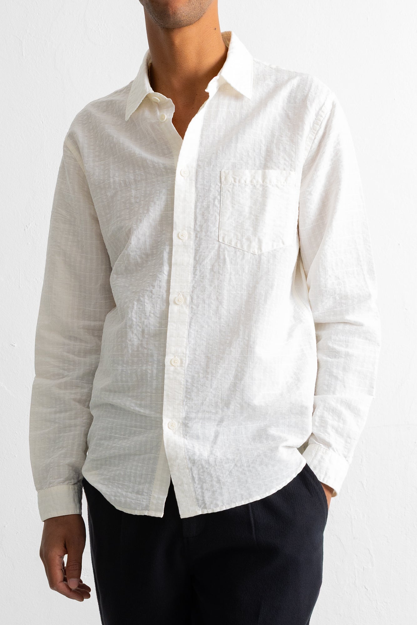 A man wearing a long-sleeve white textured linen button-down shirt with a chest pocket, paired with dark trousers, against a white wall.