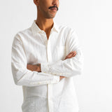 A man wearing a white long-sleeve linen button-down shirt and dark trousers, standing with arms crossed against a white wall.