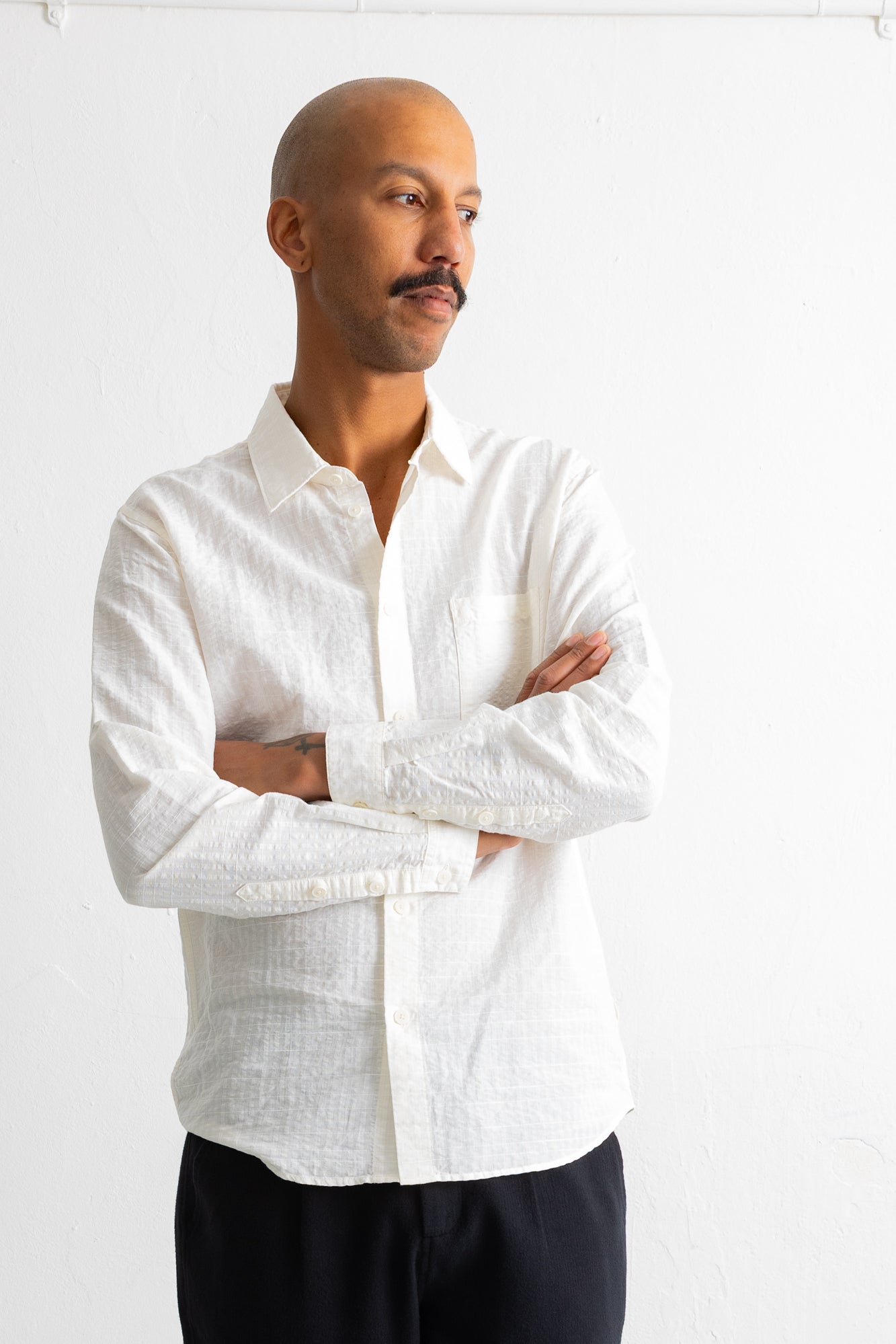 A man wearing a white long-sleeve linen button-down shirt and dark trousers, standing with arms crossed against a white wall.