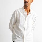 A man wearing a white long-sleeve linen button-down shirt and dark pants, standing against a white wall.