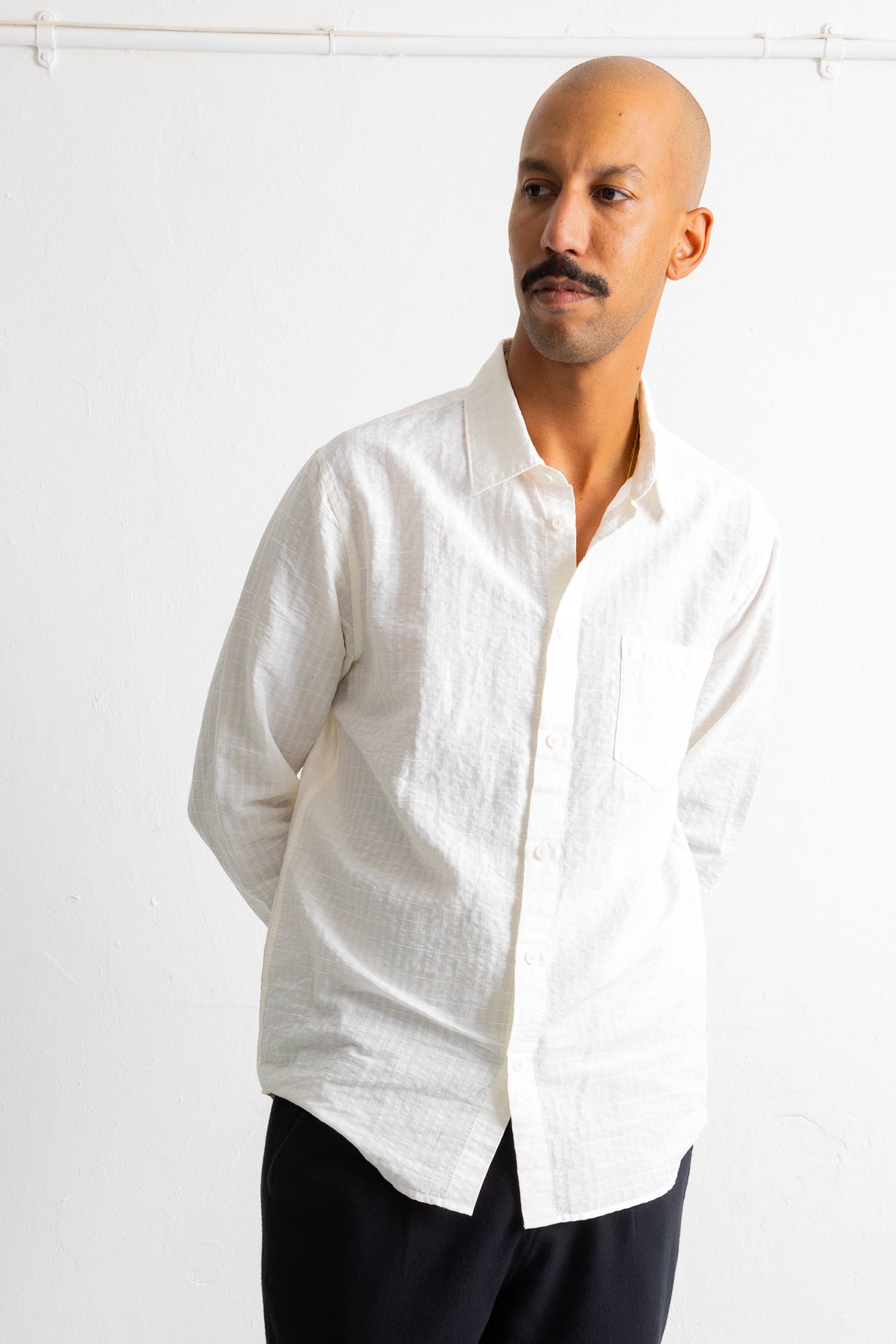 A man wearing a white long-sleeve linen button-down shirt and dark pants, standing against a white wall.