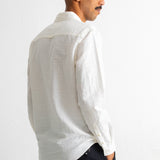 A man shown from the back and side profile wearing a white textured long-sleeve button-up shirt and dark trousers, standing against a plain white wall.