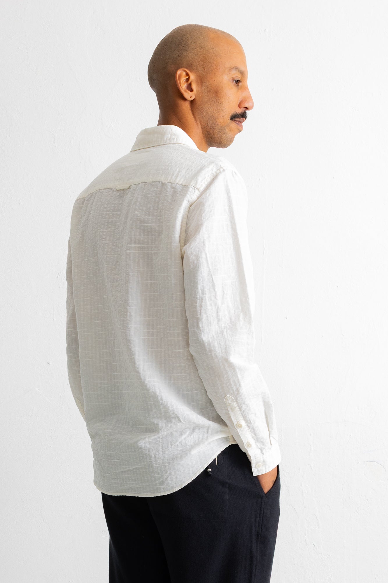 A man shown from the back and side profile wearing a white textured long-sleeve button-up shirt and dark trousers, standing against a plain white wall.