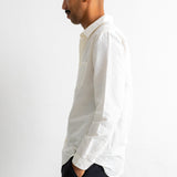 A man in profile wearing a white textured long-sleeve button-down shirt and dark trousers, standing against a plain white wall.