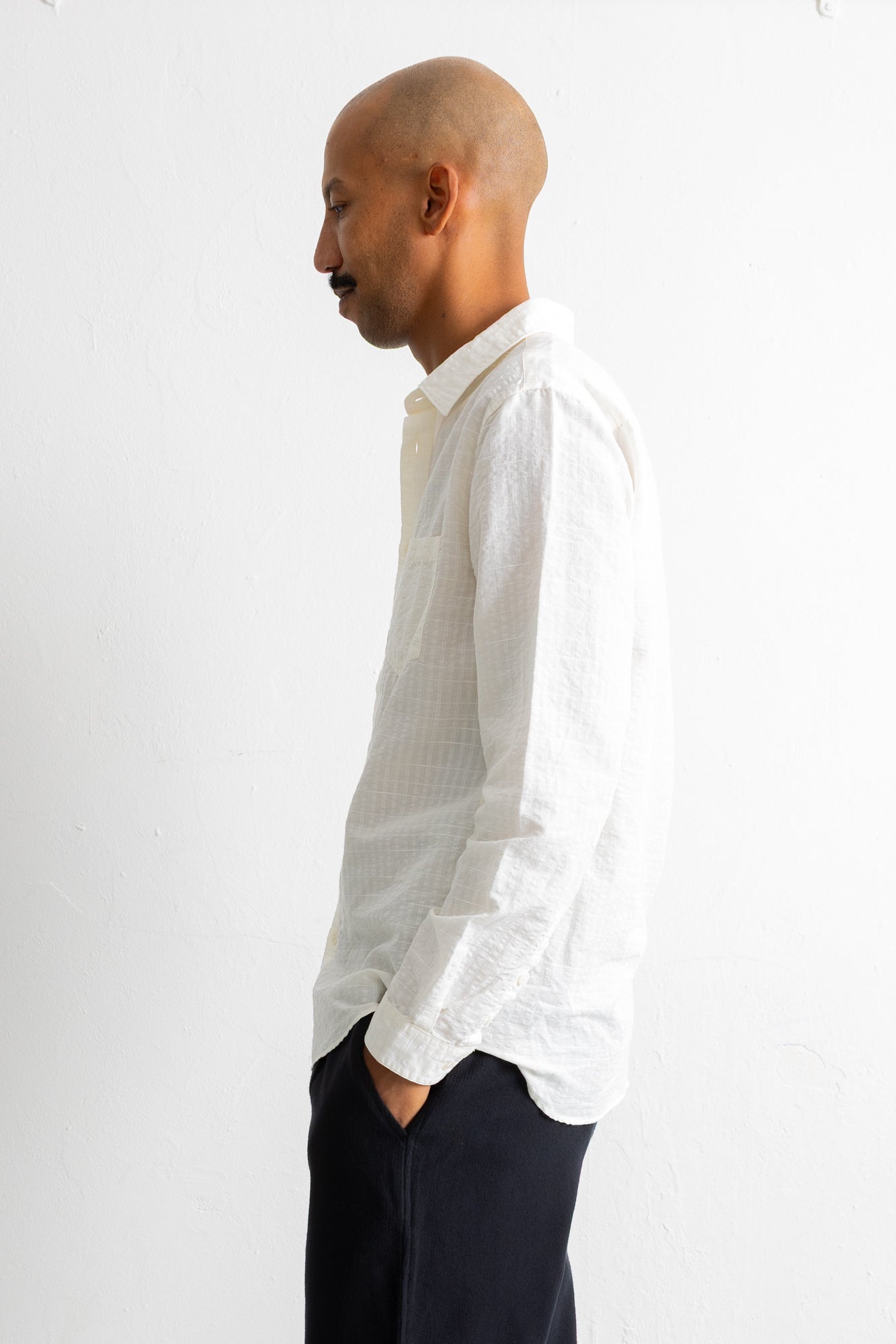A man in profile wearing a white textured long-sleeve button-down shirt and dark trousers, standing against a plain white wall.