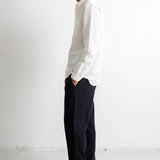 A man in a white long-sleeve button-down shirt and dark navy trousers stands in profile against a plain white wall, wearing black leather shoes.
