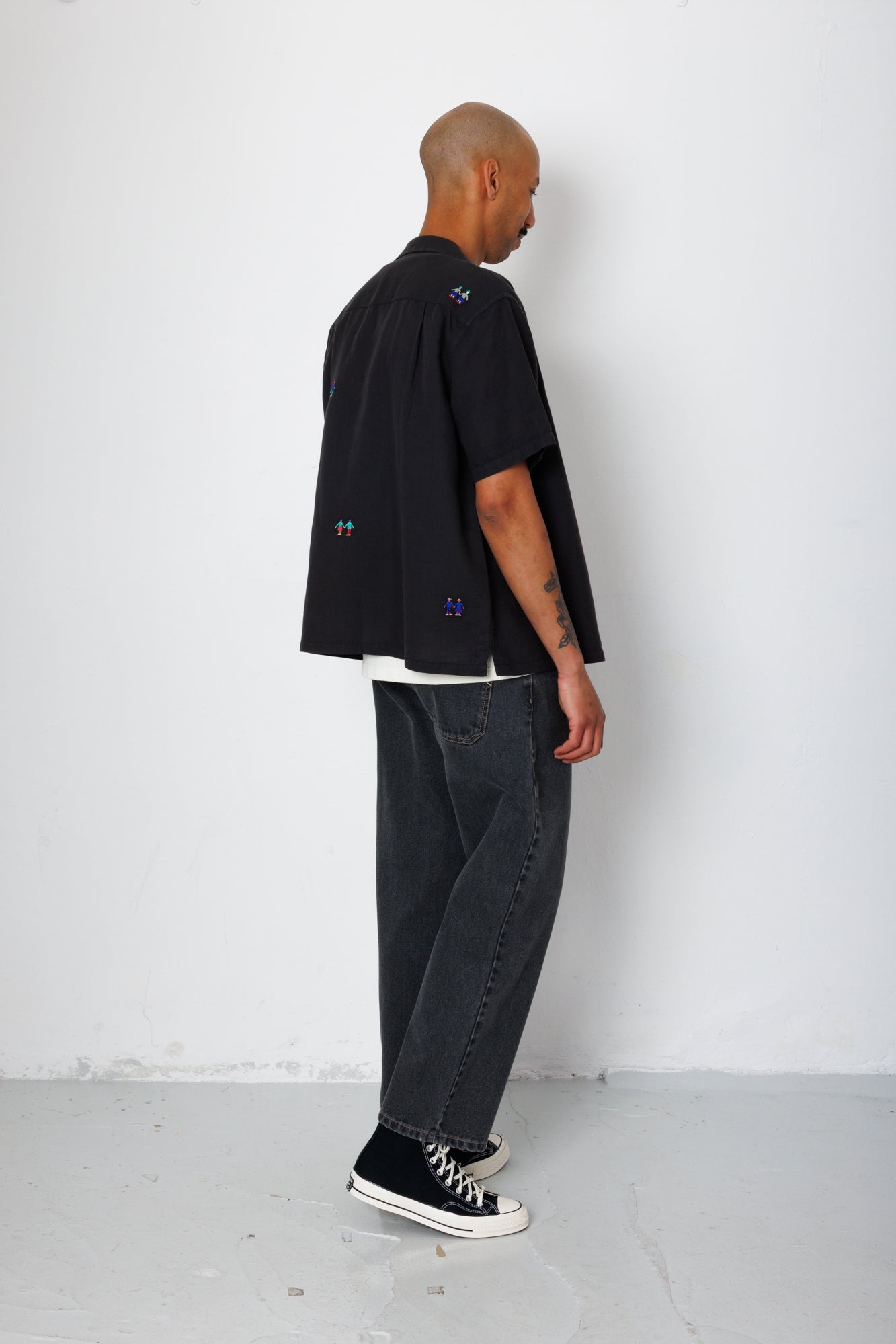Model facing a white background wearing a black shirt with embroidery. 