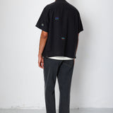 Model facing a white wall while wearing a black shirt with embroidery.